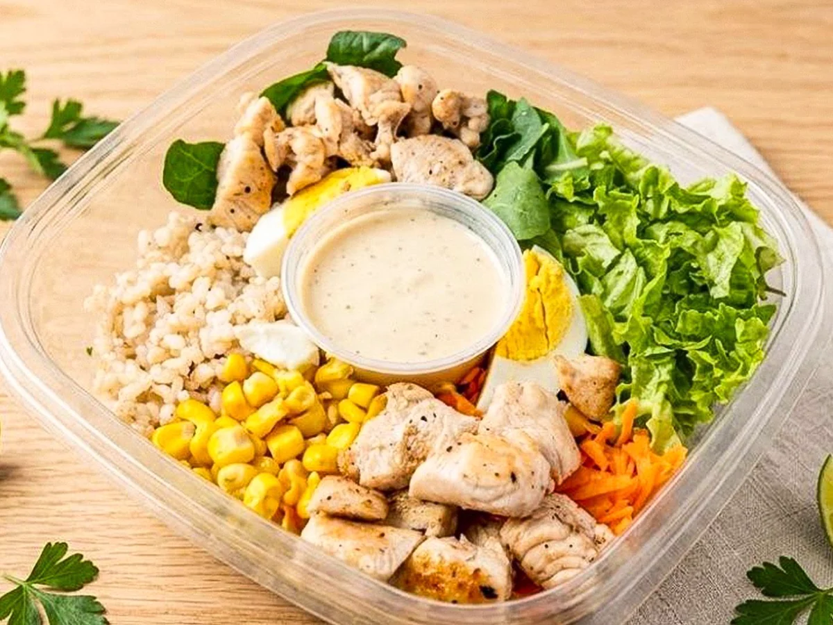 Ensalada con pollo, hojas verdes, huevo, maíz, zanahoria y un pote de aderezo.